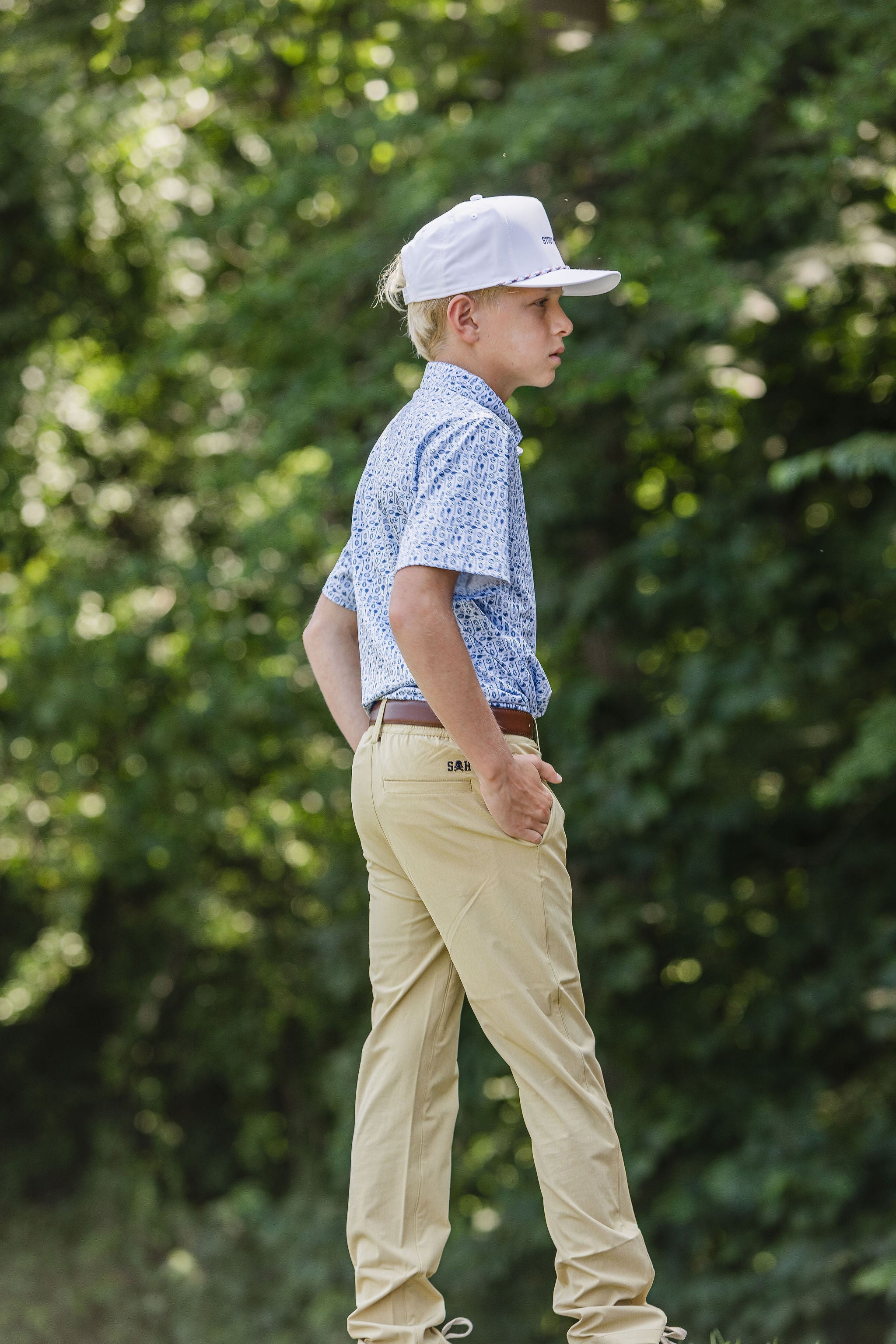 Young kid wearing long pant khakis, blue pattern polo top, and white hat in front of greenery background