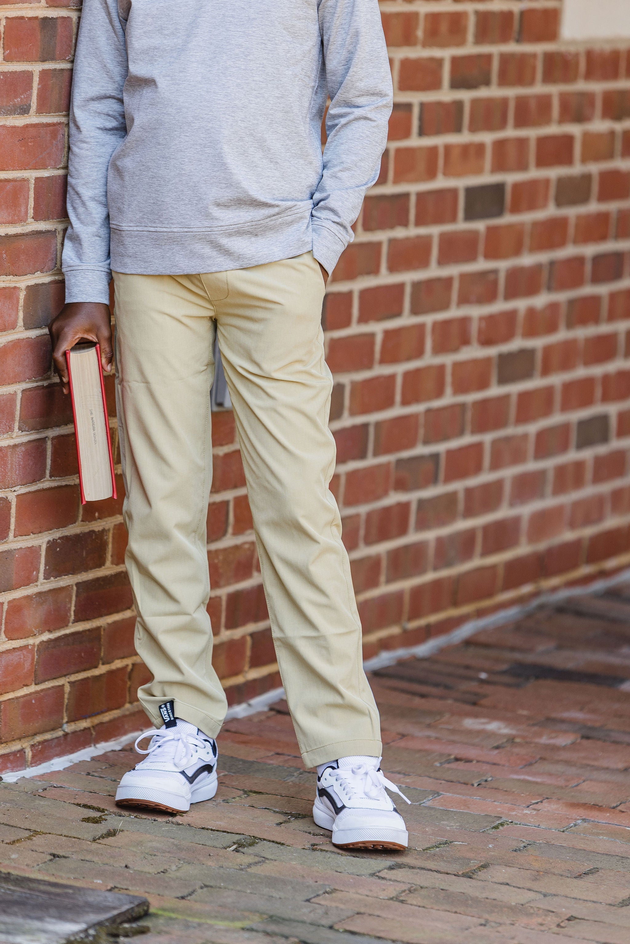 Young person leaning against brick wall holding book wearing white tennis shoes, long pant khakis, and gray quarter zip