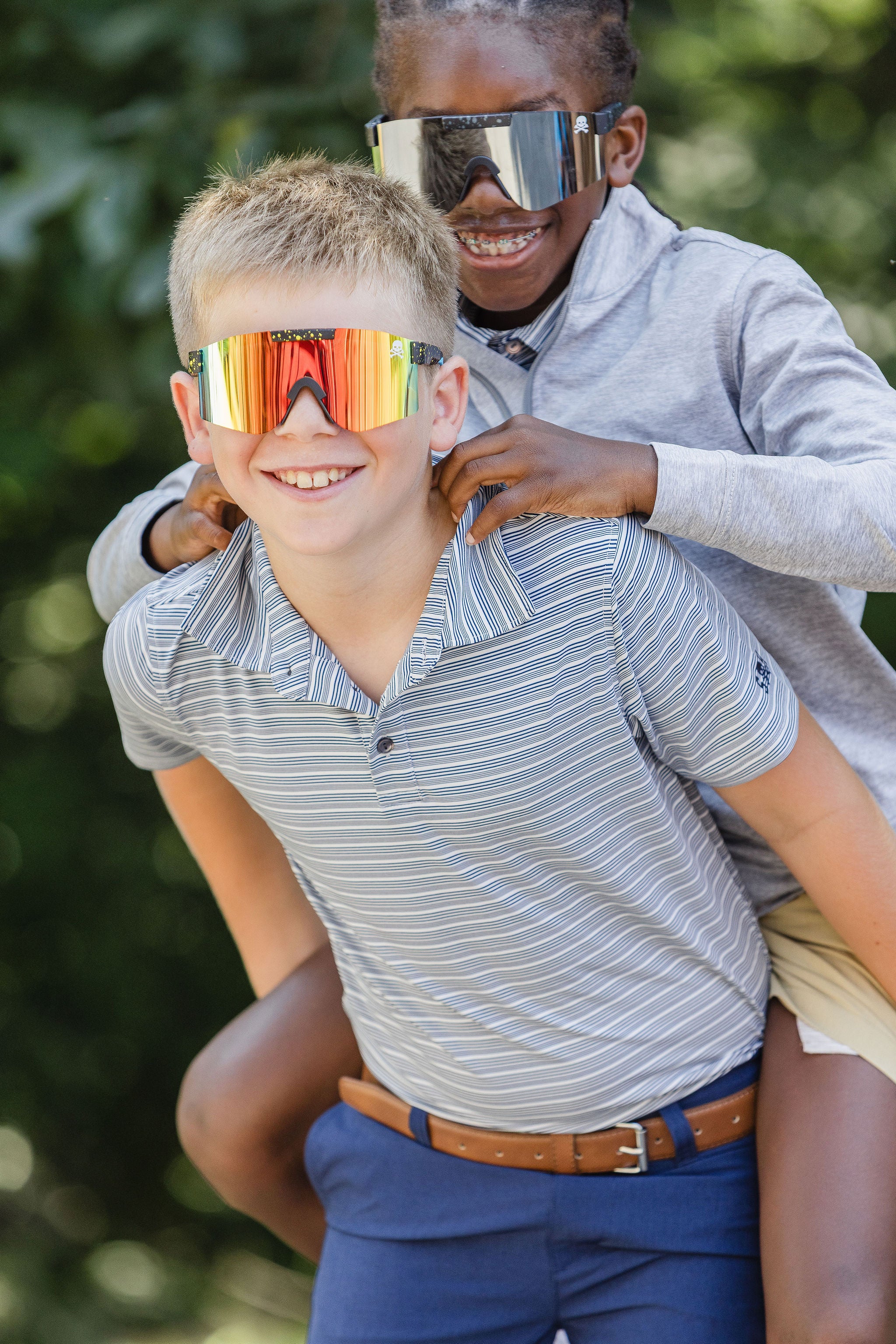 Two children wearing colorful reflective sunglasses outdoors with a blurred green background