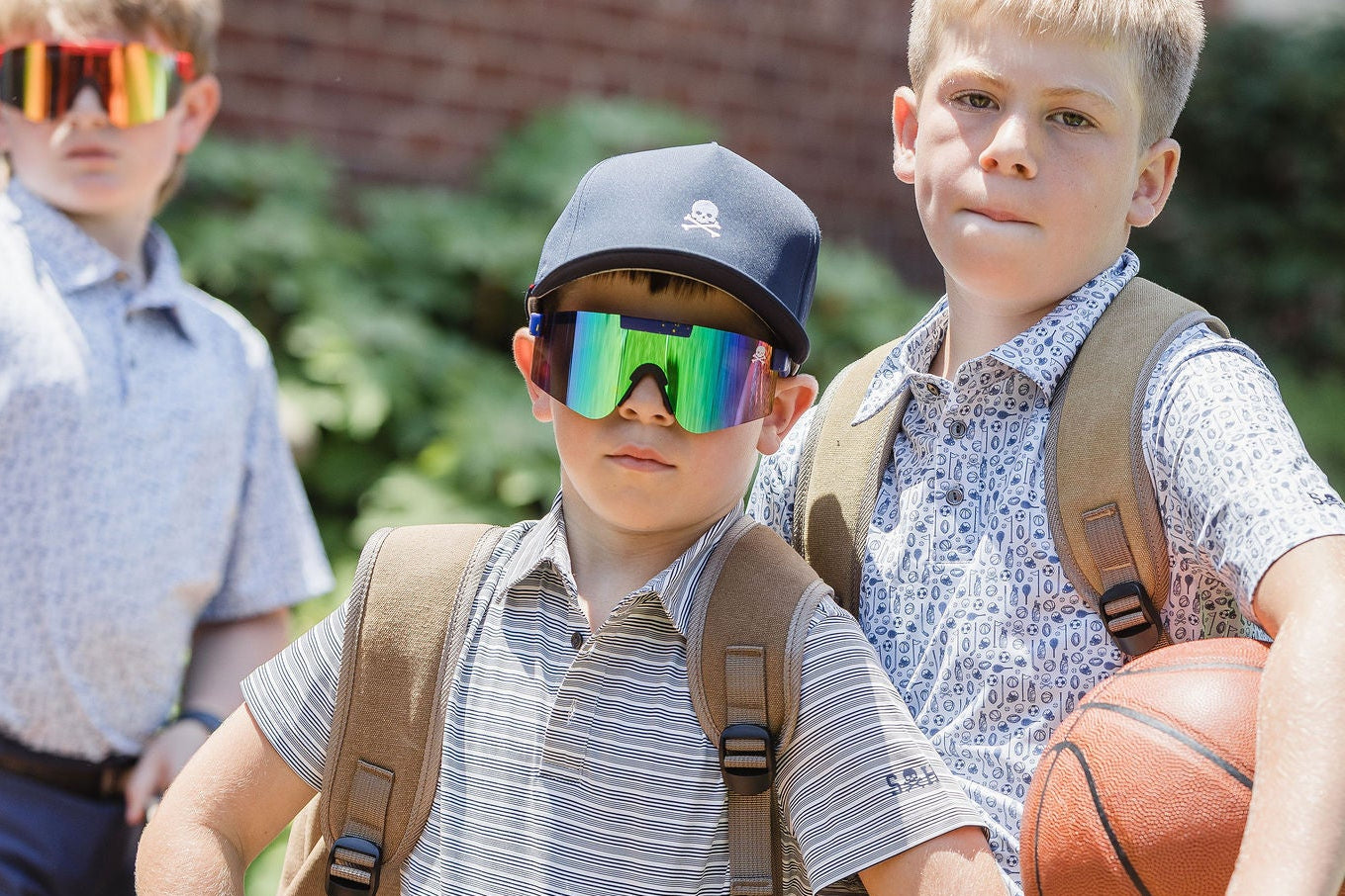 Two boys with backpacks, one wearing sunglasses, and holding a basketball, standing outdoors