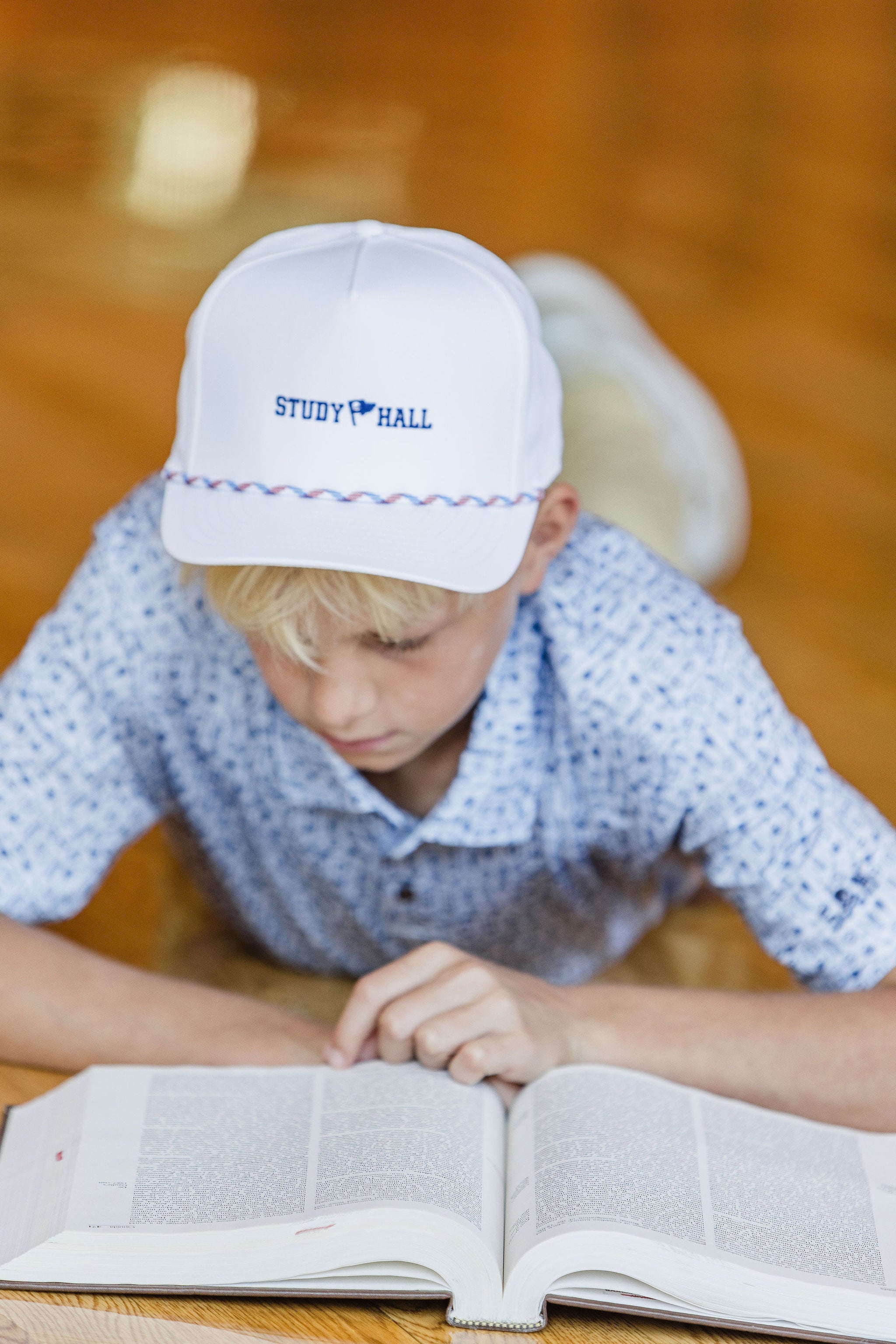 Child wearing a 'Study Hall' cap reading a book on a wooden table.