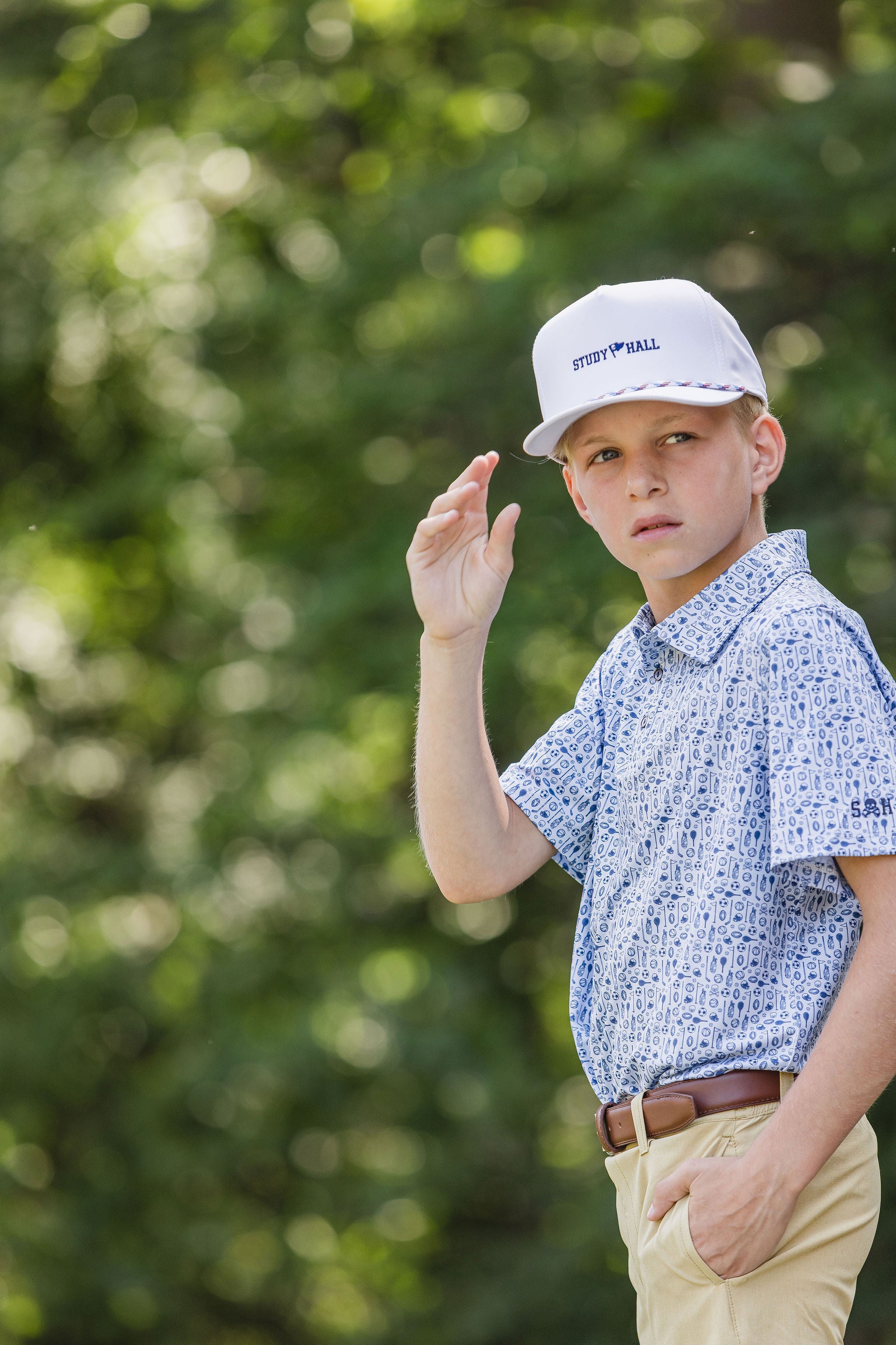 Young boy wearing a white cap and patterned shirt outdoors with greenery in the background