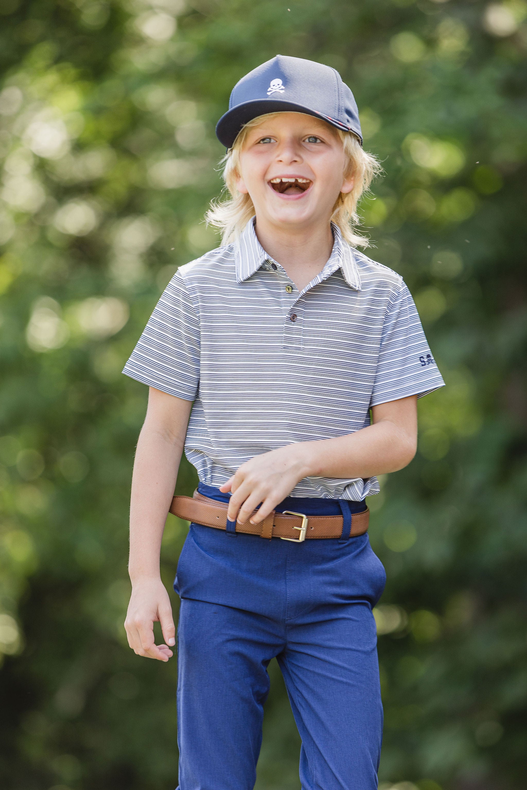 Young boy in a striped shirt and blue pants standing outdoors with a blurred green background wearing navy hat