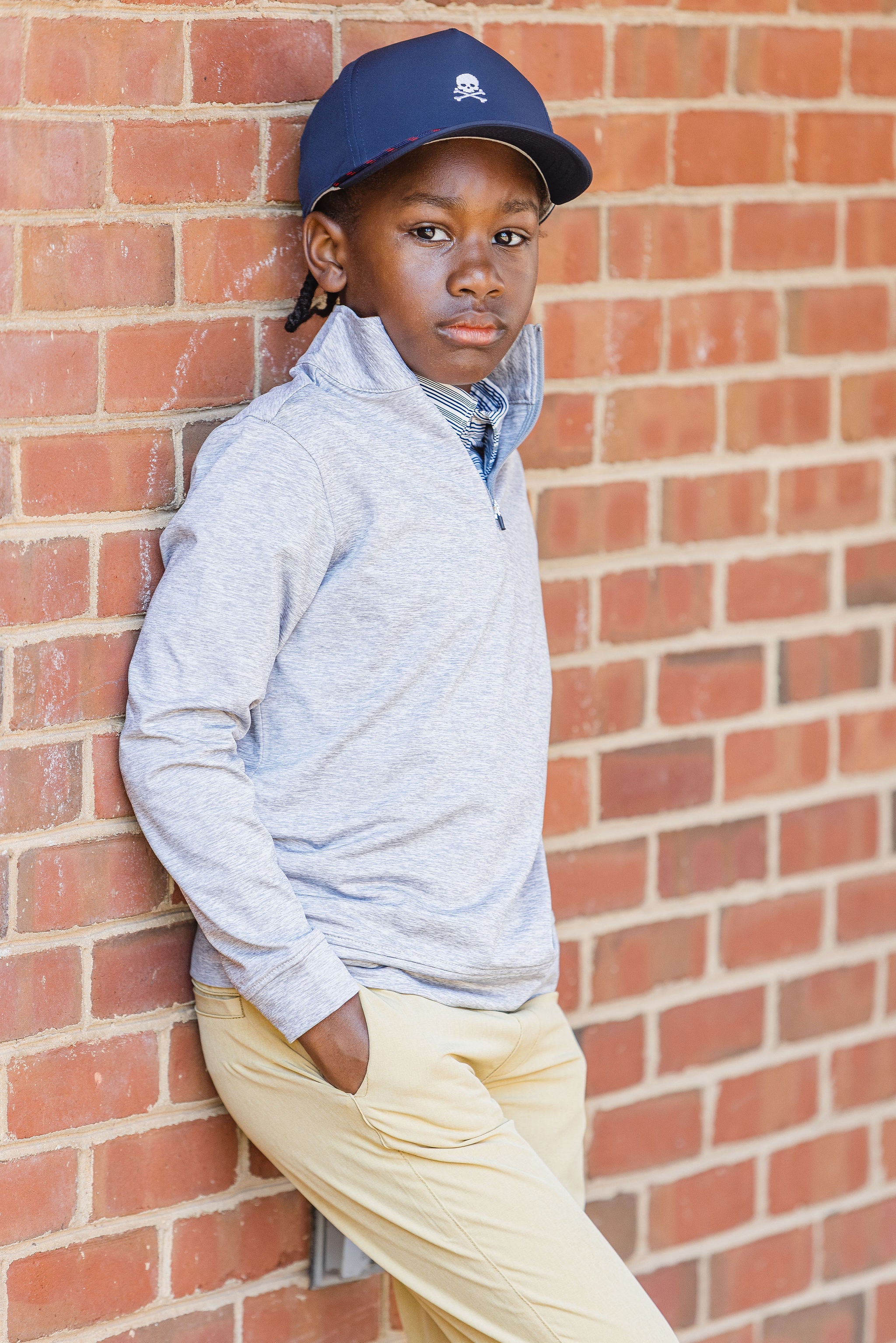 Young boy wearing navy cap, gray quarter zip, and long pant khakis leaning on brick wall