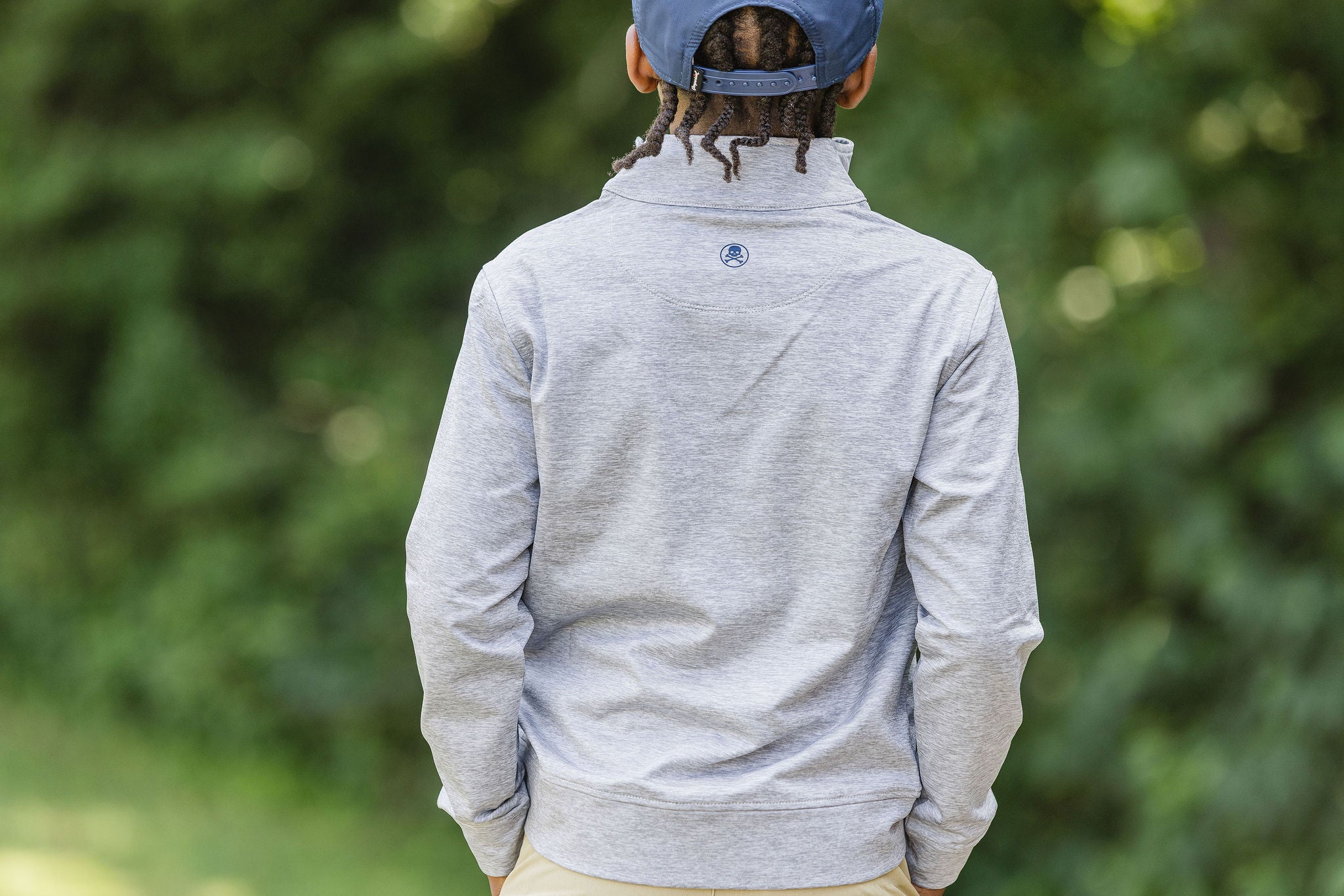 Person wearing a gray zip-up hoodie with a logo on the back, standing outdoors with greenery in the background.
