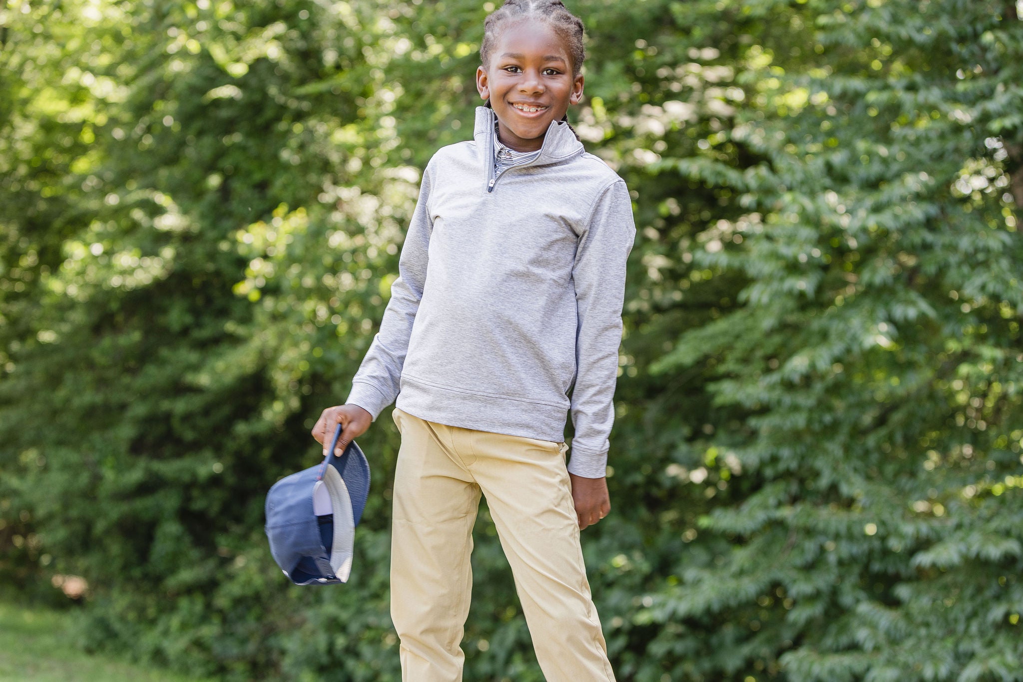 Child wearing a gray quarter zip jacket and beige pants, holding a blue cap, with green trees in the background