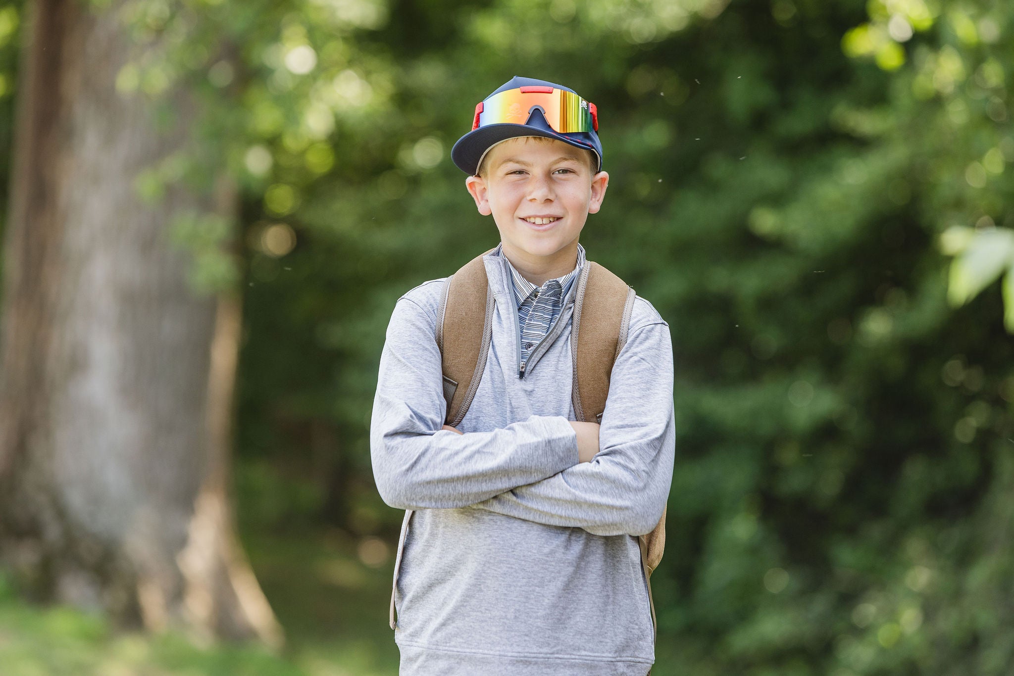 Young boy wearing cap, sunglasses, and backpack standing outdoors with greenery in the background