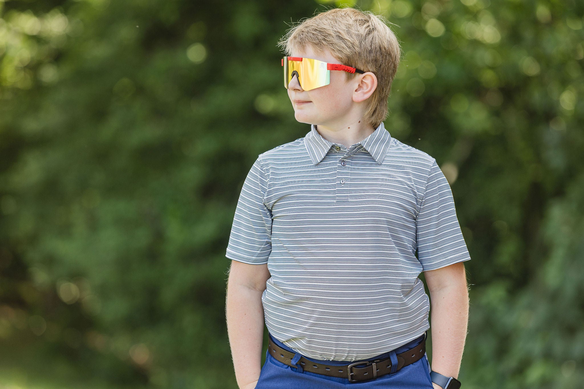 Young boy wearing a striped polo shirt and reflective sunglasses outdoors.