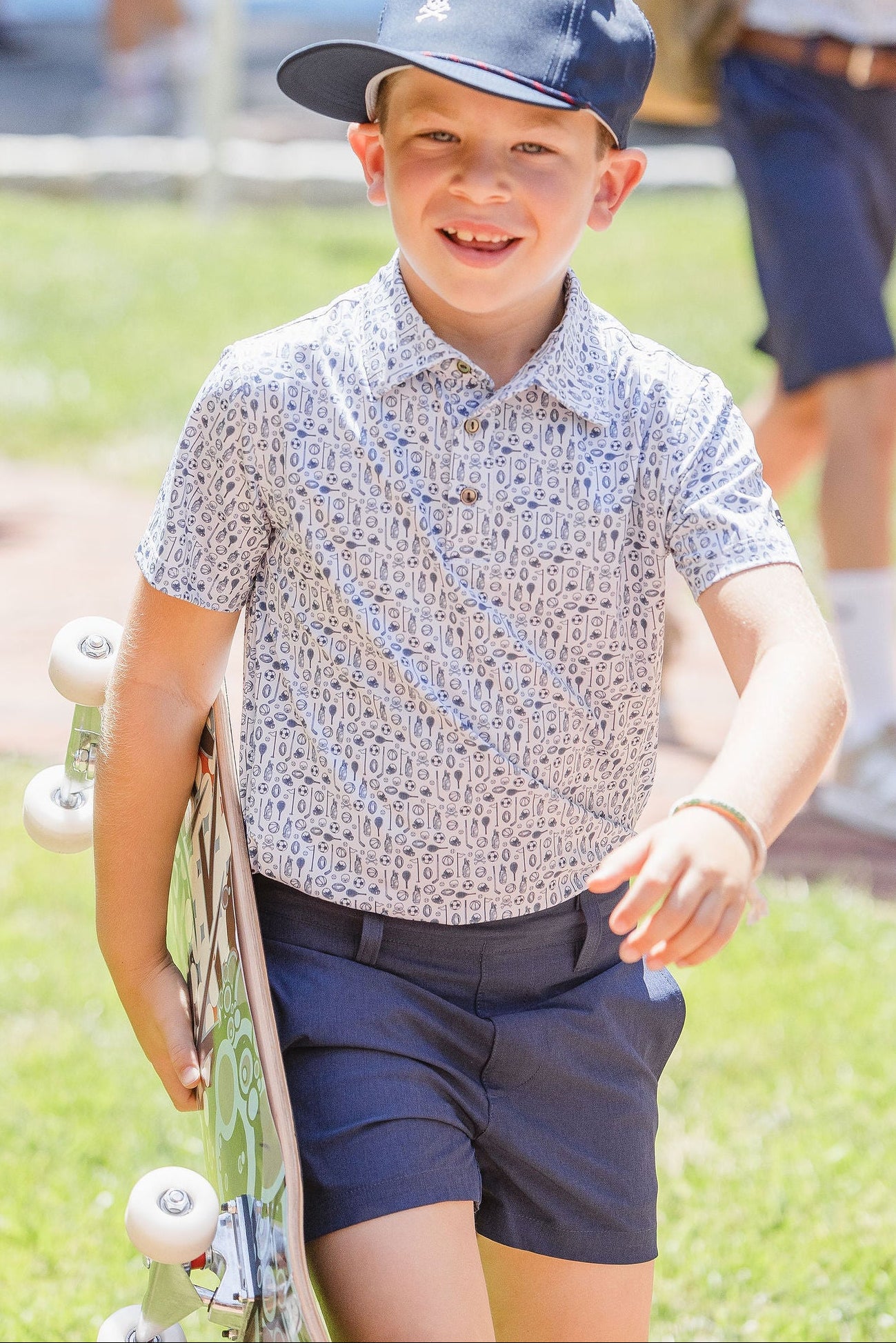 Boy wearing sideways navy ball cap and pattern polo carrying skateboard at park