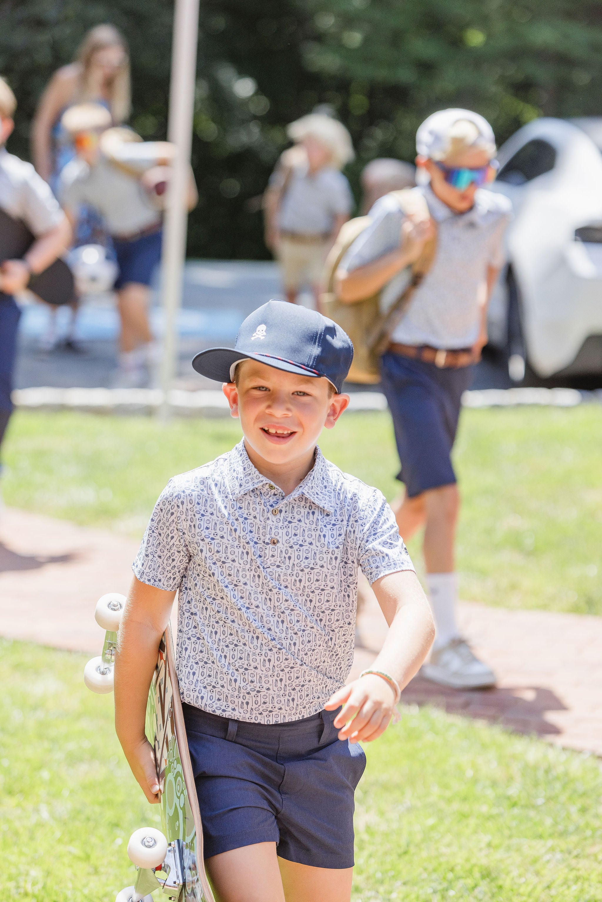 Boy wearing sideways navy ball cap and pattern polo carrying skateboard at park