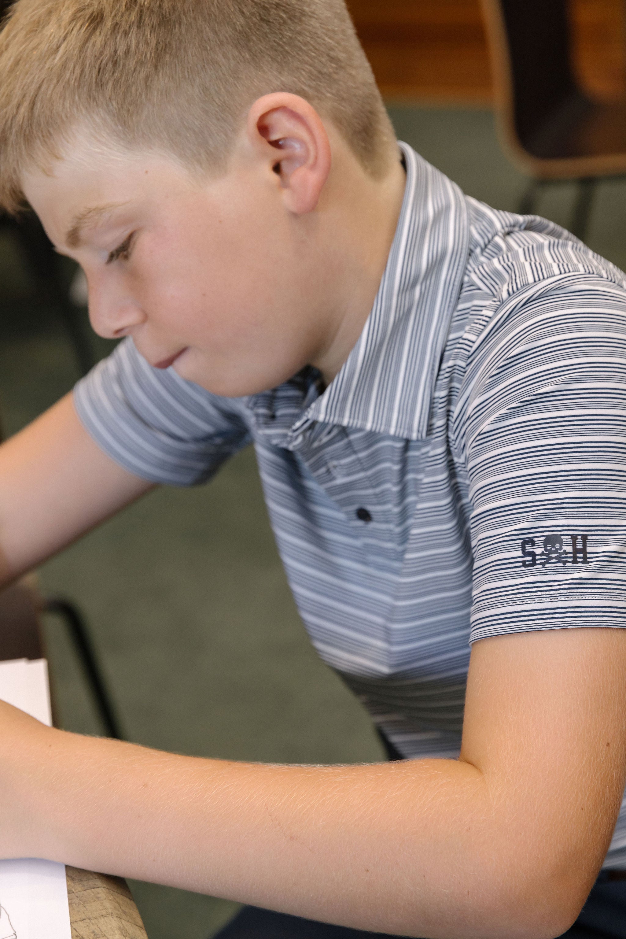 Young boy wearing a striped polo shirt with a logo, sitting and looking down.
