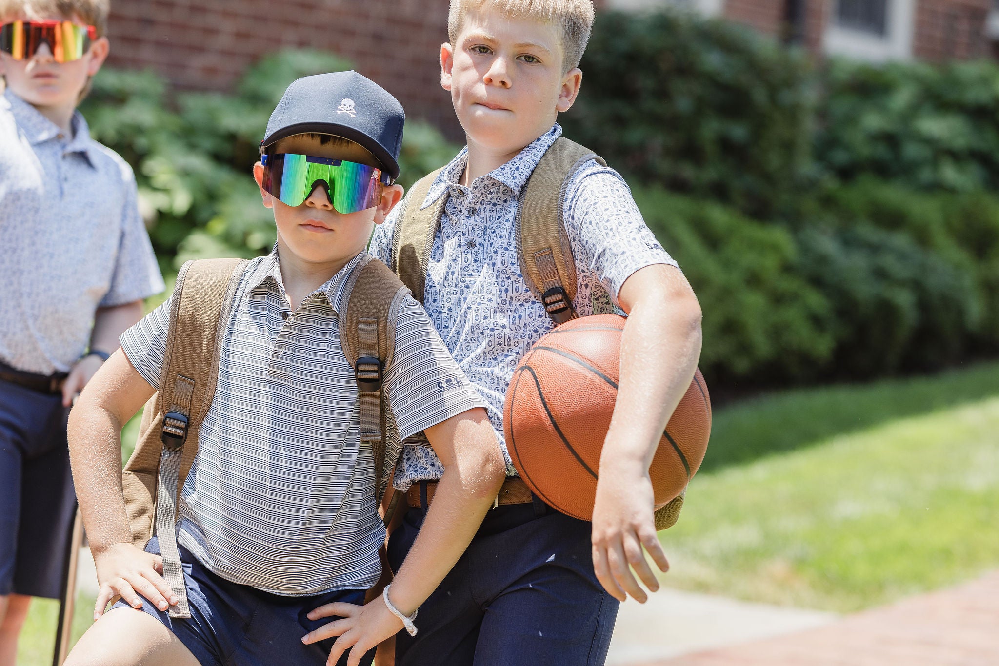 Two boys with backpacks, one wearing sunglasses, and holding a basketball, standing outdoors