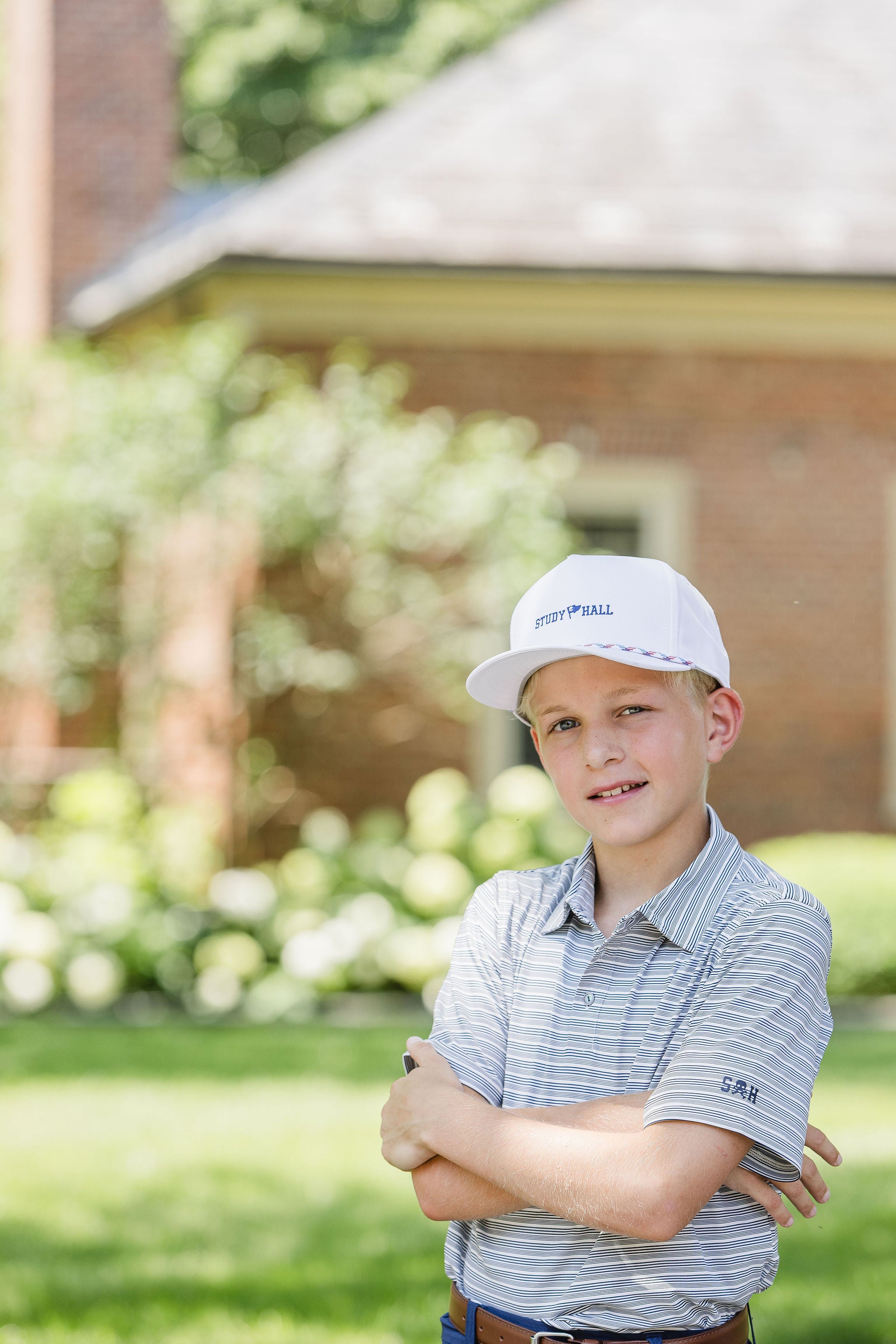 Young boy wearing a white cap and striped shirt standing outdoors with a blurred school and garden in the background.